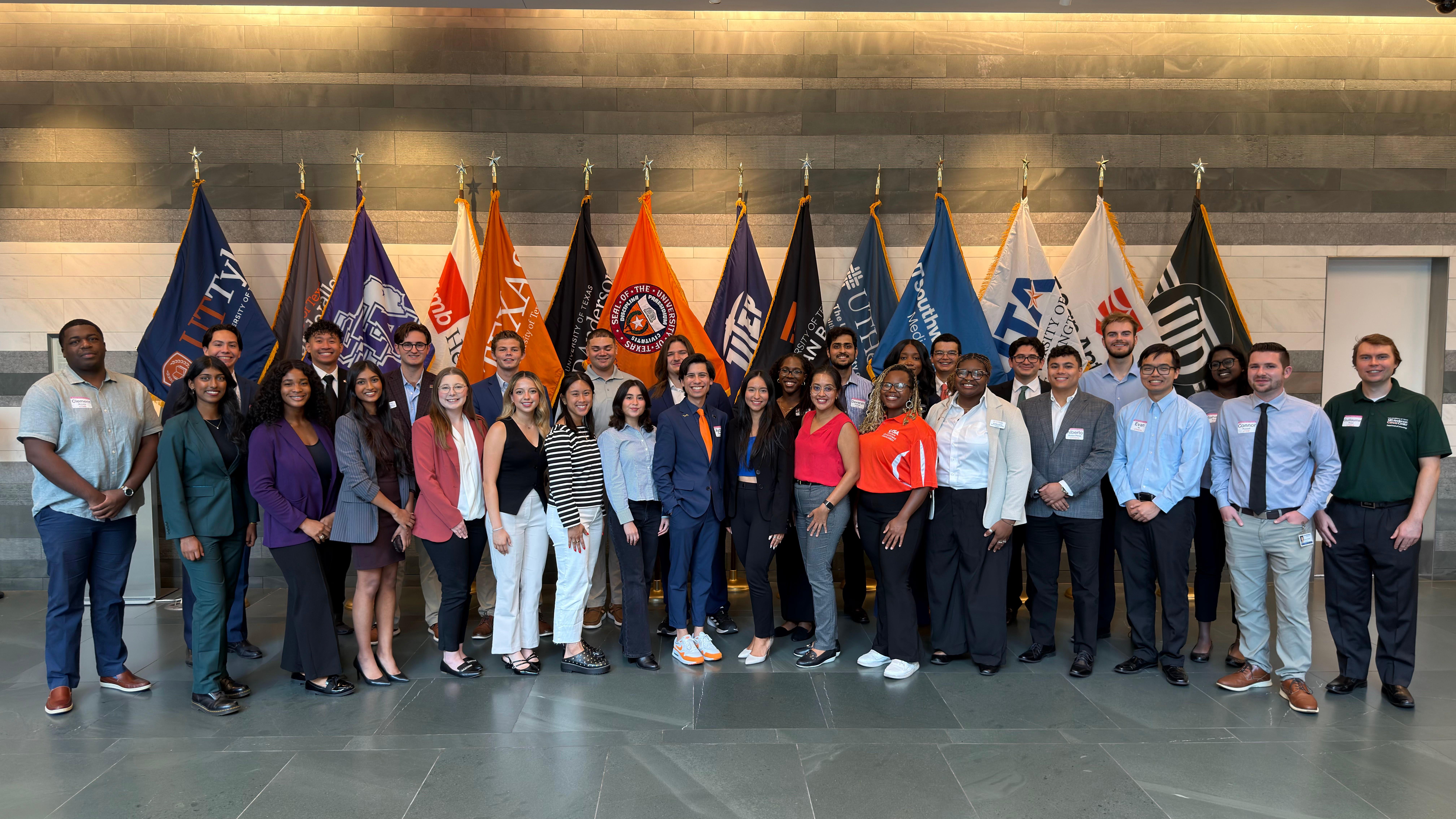 Large group photo of students and staff standing indoors in front of university flags during a meeting event